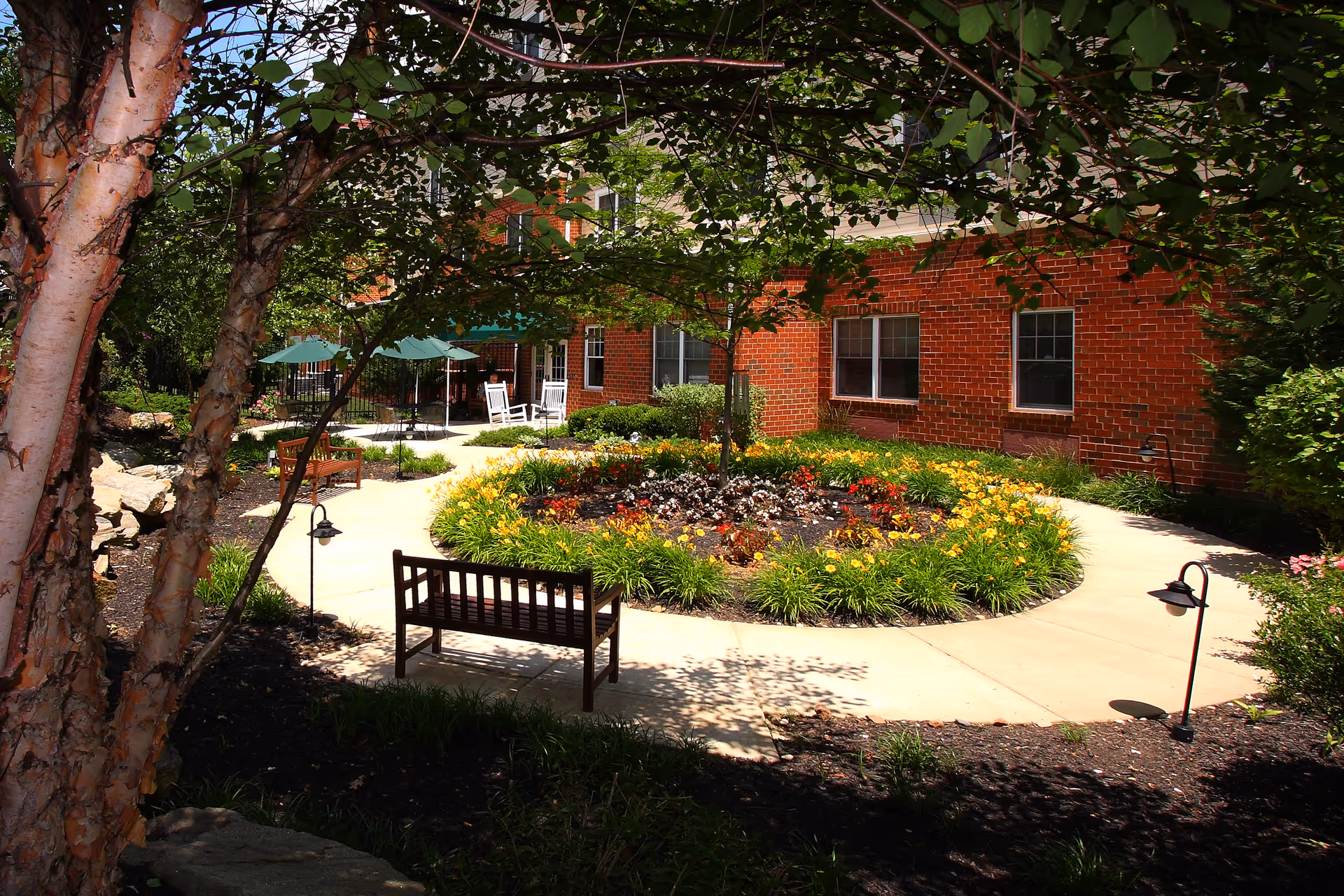 A peaceful outdoor garden area with a circular flower bed surrounded by a concrete walkway. There are wooden benches, green umbrellas, and white rocking chairs near a red brick building. Trees and shrubs provide shade and greenery.