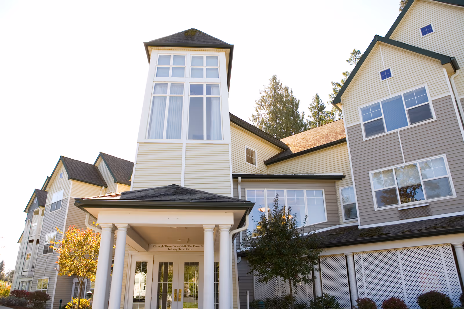 Exterior view of a multi-story senior living facility building with beige and gray siding, large windows, a covered entrance supported by white columns, and surrounding trees and shrubs.