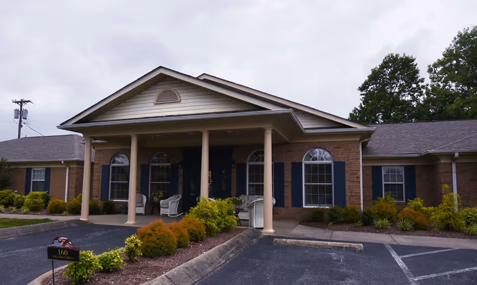 Front exterior view of a single-story brick building with a covered entrance supported by four columns. The building has multiple windows with dark shutters and a small landscaped area with bushes and plants in front. There are a few white chairs on the porch and a parking area in front.