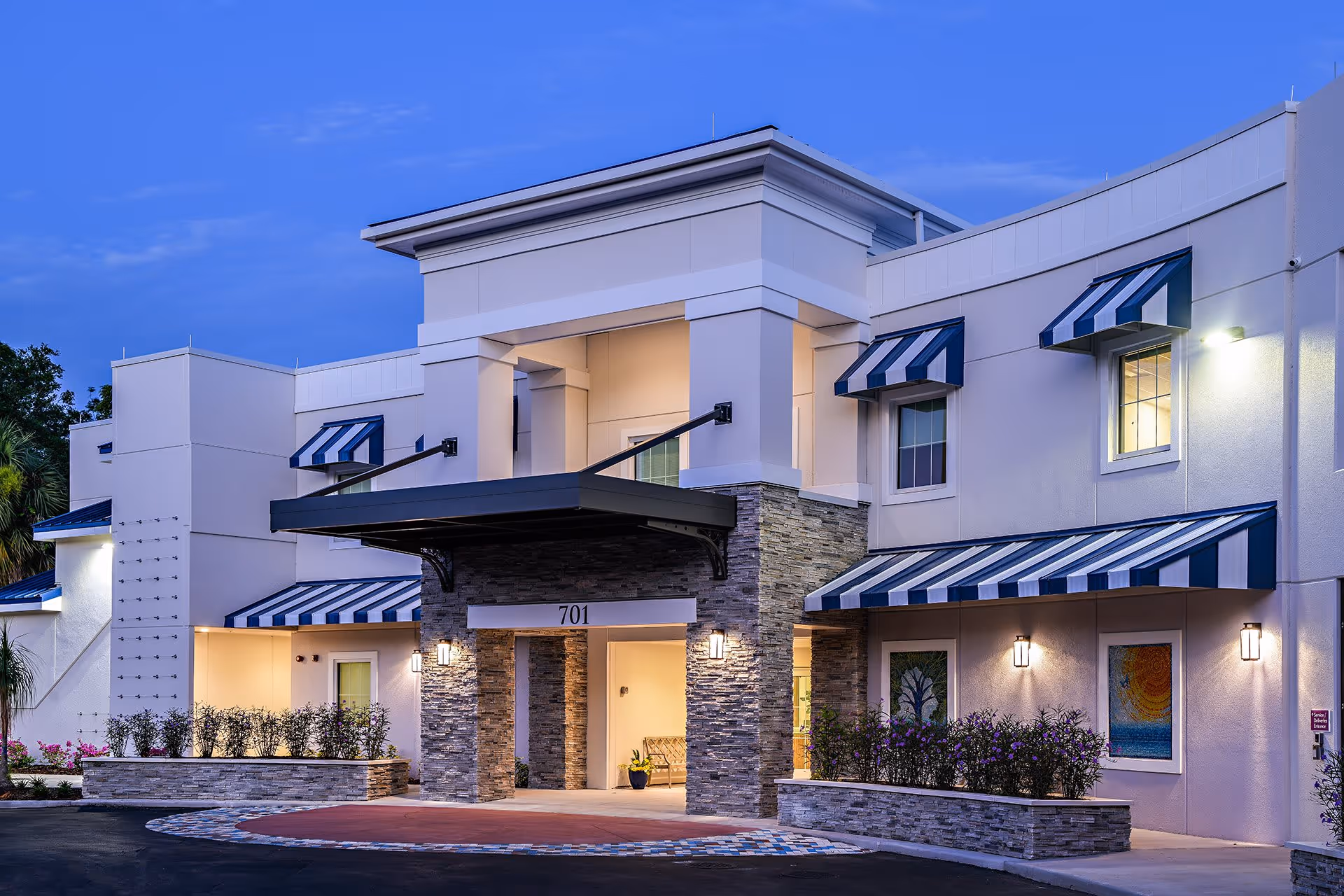 Exterior view of a modern senior living facility building at dusk with stone pillars, striped blue and white awnings over windows and entrance, and well-lit entrance area with the number 701 displayed above the doorway.