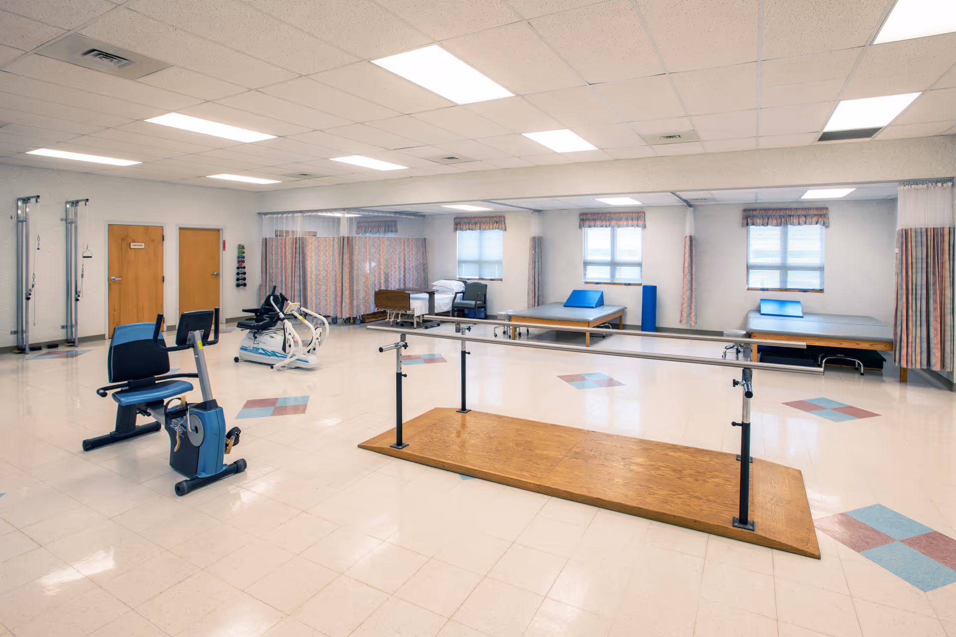 A spacious rehabilitation room with exercise equipment including stationary bikes, parallel bars for walking practice, and therapy beds along the back wall. The room has tiled floors, fluorescent ceiling lights, and windows with curtains. There are also privacy curtains and doors on the left side of the room.