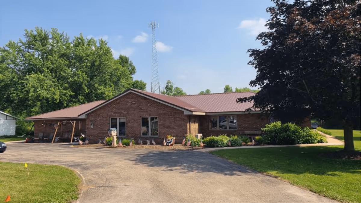Single-story brick building with a brown metal roof surrounded by green trees and bushes under a blue sky with some clouds. There is a paved driveway and a grassy lawn in front of the building.