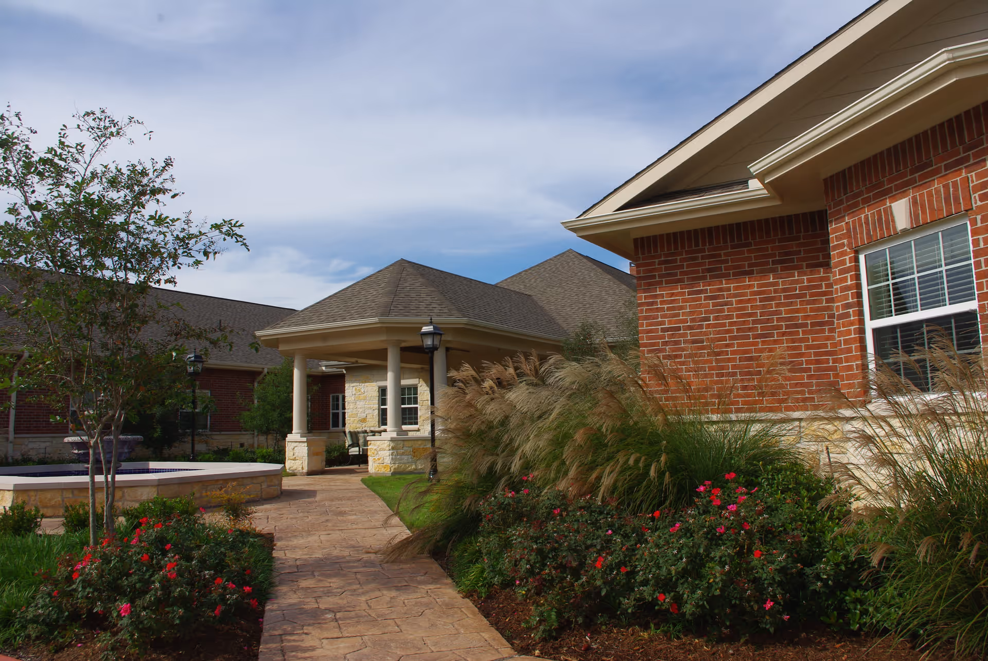 Outdoor walkway at The Heritage of Meyerland facility with brick buildings on either side, landscaped garden beds with bushes and flowers, a tree, and a covered entrance supported by columns under a partly cloudy sky.