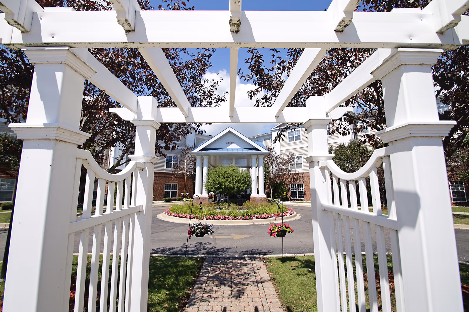 View through a white wooden pergola gate looking towards the entrance of a senior living facility building with a circular driveway, landscaped flower beds, and trees under a blue sky.