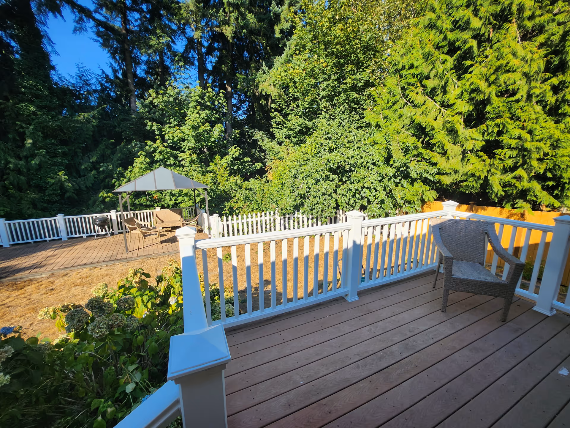 Wooden deck with white railing and a chair overlooking a yard with a gazebo and dense trees.