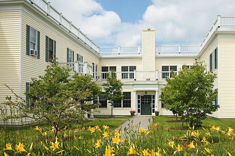 Exterior view of a light yellow two-story building with white trim, green doors, and multiple windows. The building surrounds a courtyard with green grass, small trees, and yellow flowers in the foreground under a partly cloudy sky.