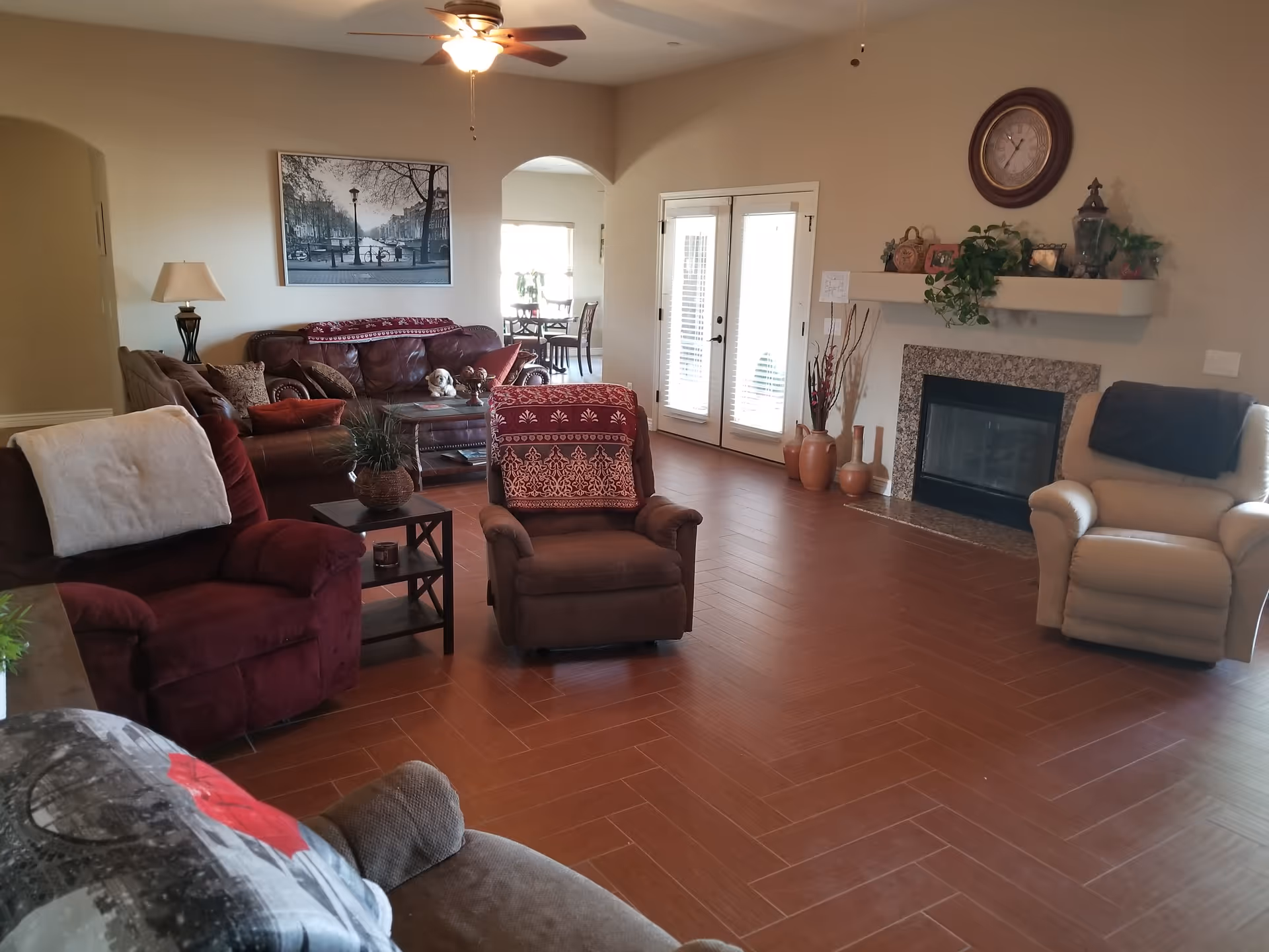 A cozy living room with multiple armchairs and sofas arranged around a coffee table. The room features a ceiling fan with a light, a fireplace with decorative items on the mantel, a wall clock above the fireplace, and French doors letting in natural light. There is a framed black and white photo on the wall and a small side table with a lamp.