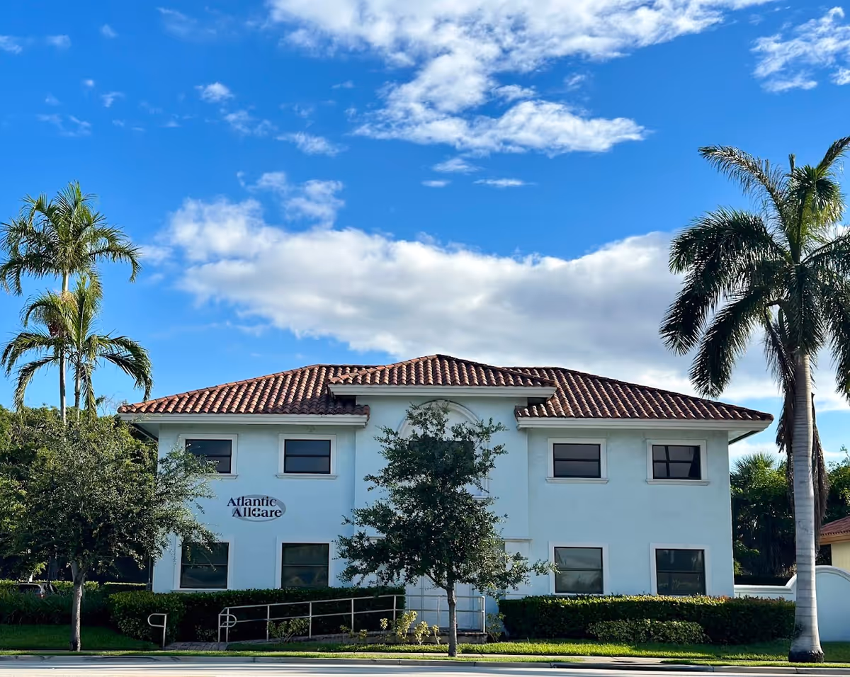 White two-story building with red-tiled roof, palm trees, and Atlantic AllCare sign.