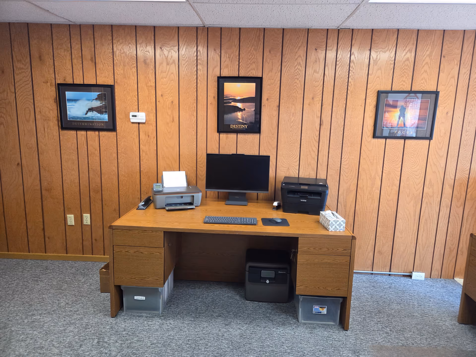 Wood-paneled office with desk, computer, printer, and framed posters on walls.