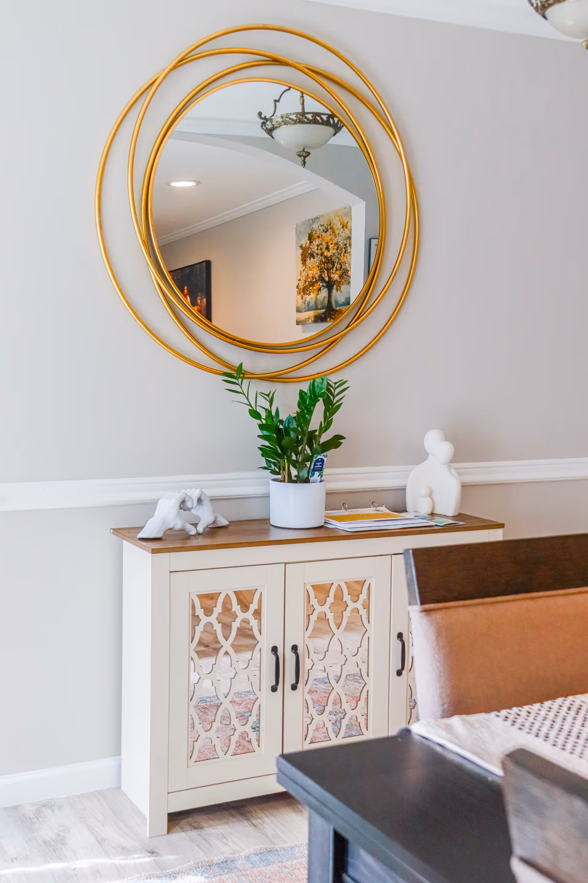 White cabinet with lattice doors, circular gold mirror, plant, and seating in a modern dining area.