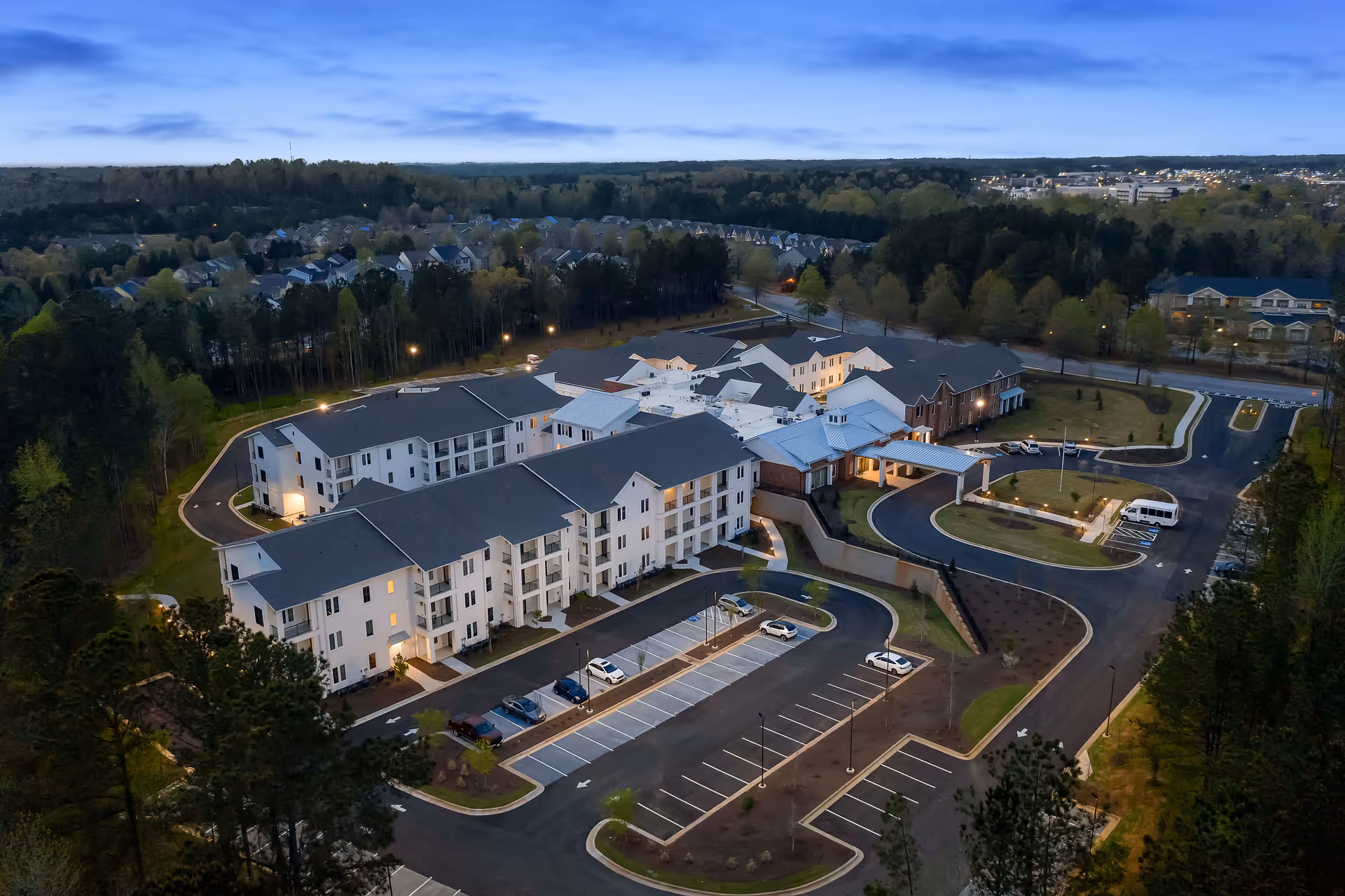 Aerial evening view of a multi-building senior living campus with lit walkways, parking lots, and surrounding trees.