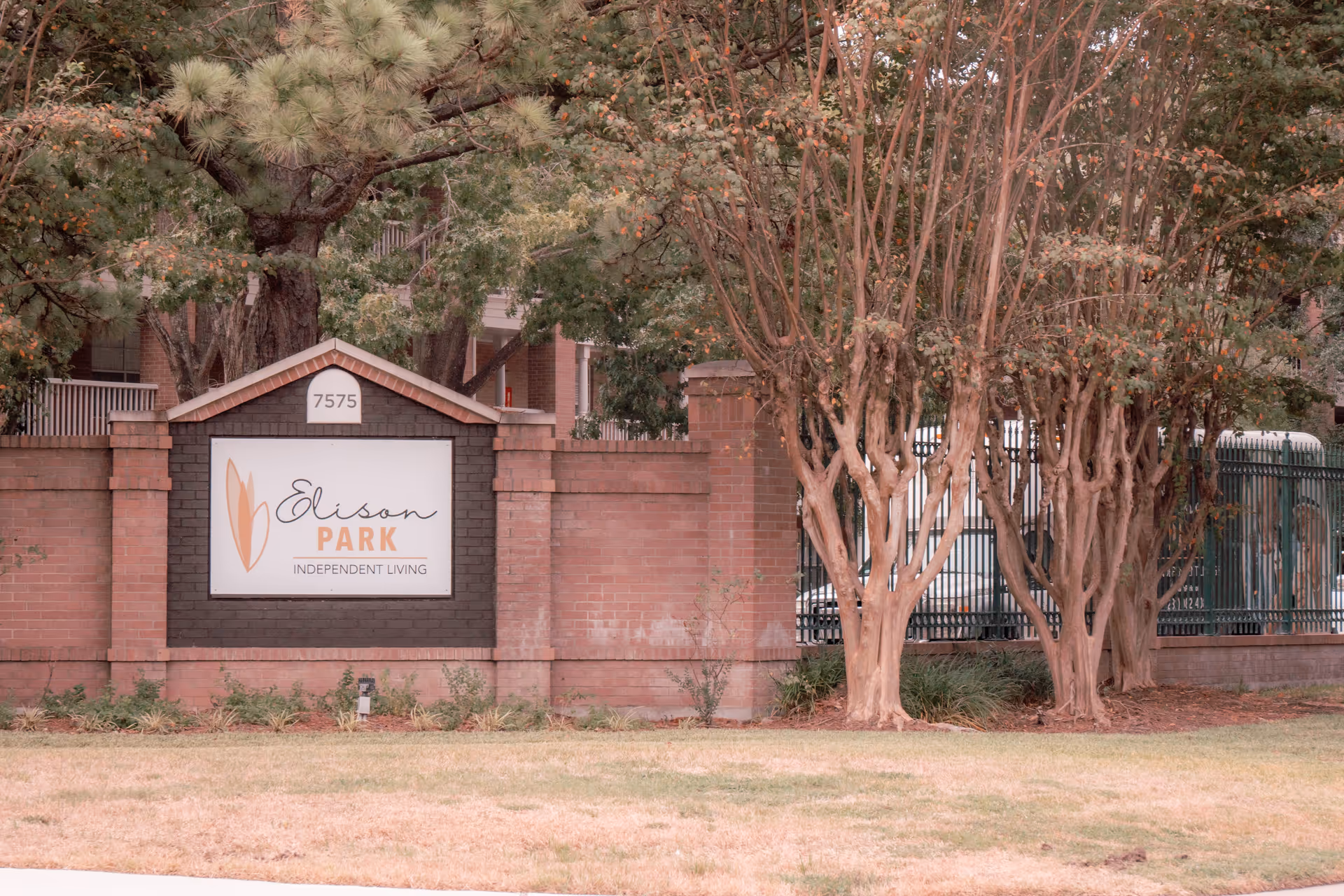 A brick wall with a sign that reads 'Elison Park Independent Living' surrounded by trees and greenery, with a building partially visible in the background.