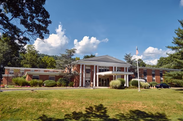 Red brick senior care building with columns, entrance, flagpole, and manicured lawn.