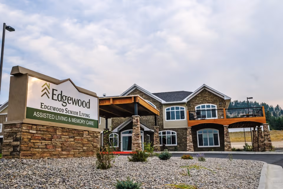 Exterior view of Edgewood Helena senior living facility showing a two-story building with stone and wood accents, large windows, and a covered entrance. In the foreground, there is a large sign that reads 'Edgewood Senior Living Assisted Living & Memory Care' surrounded by landscaping with rocks and small plants.
