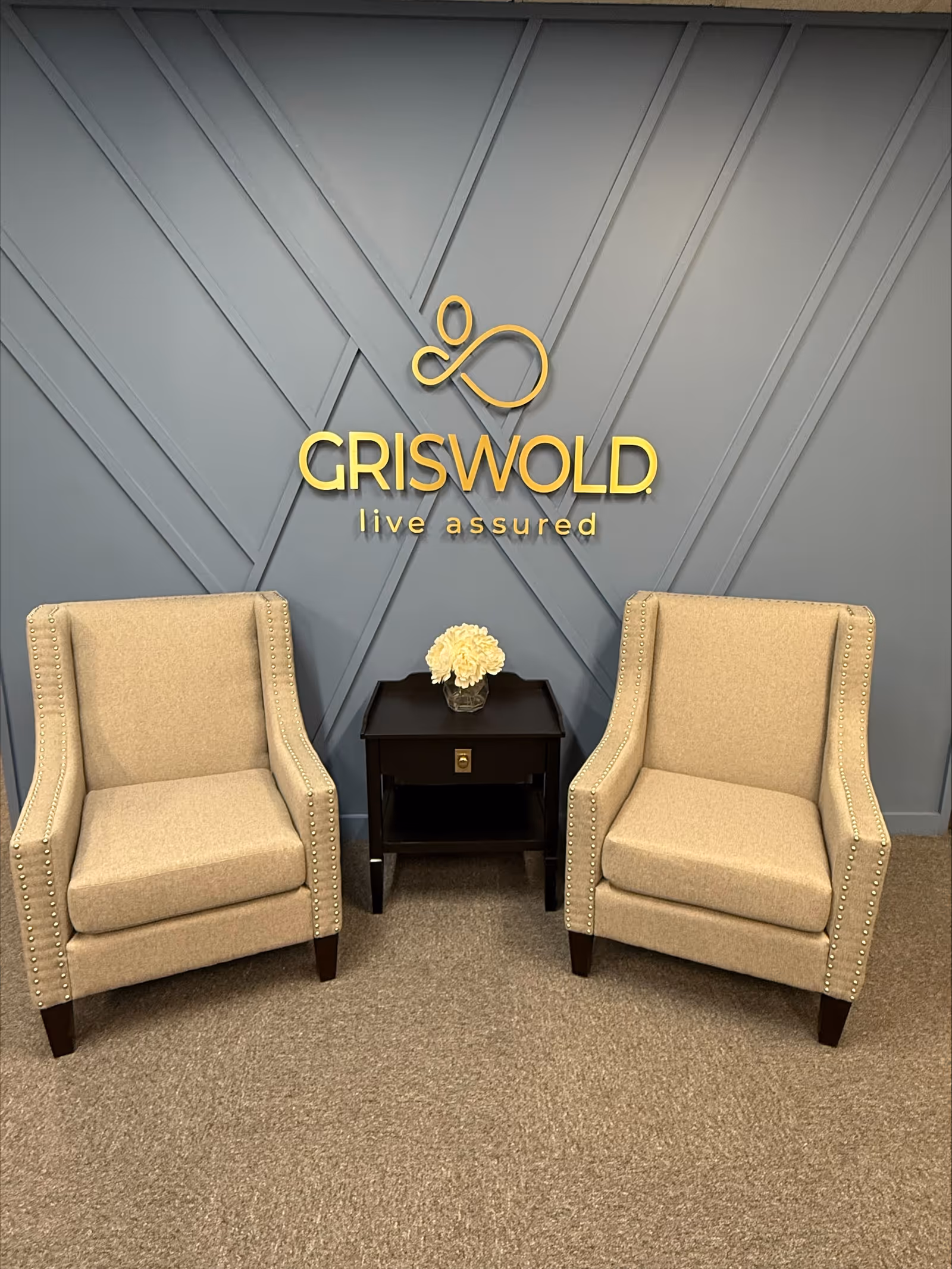 Two beige armchairs flank a side table with a flower arrangement against a blue paneled wall.