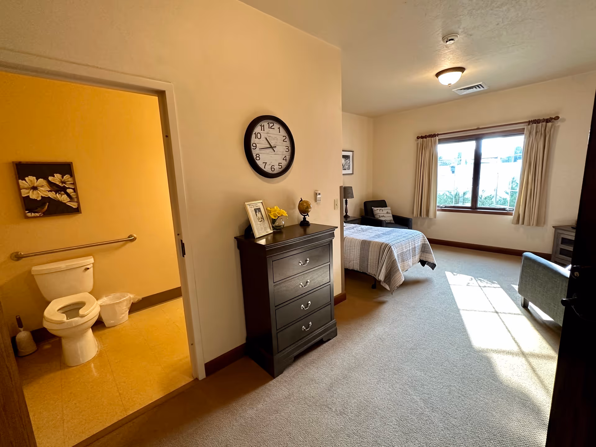 Bedroom in a senior living suite with bed, dresser, chair, and window.