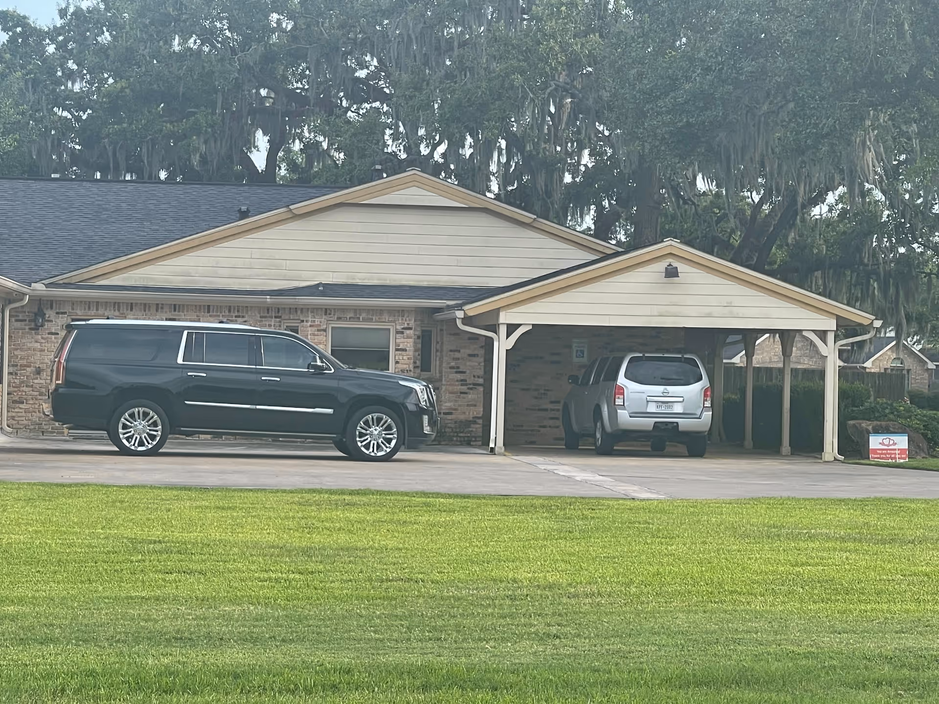 Single-story brick assisted living building with a covered carport and two parked SUVs in front of a green lawn.