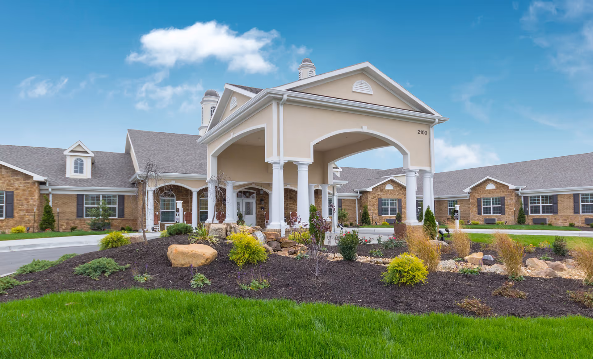 Front exterior view of Benton House of Raymore, featuring a large covered entrance with white columns, beige walls, and stone accents. The building is surrounded by landscaped gardens with green grass, shrubs, and rocks under a blue sky with some clouds.