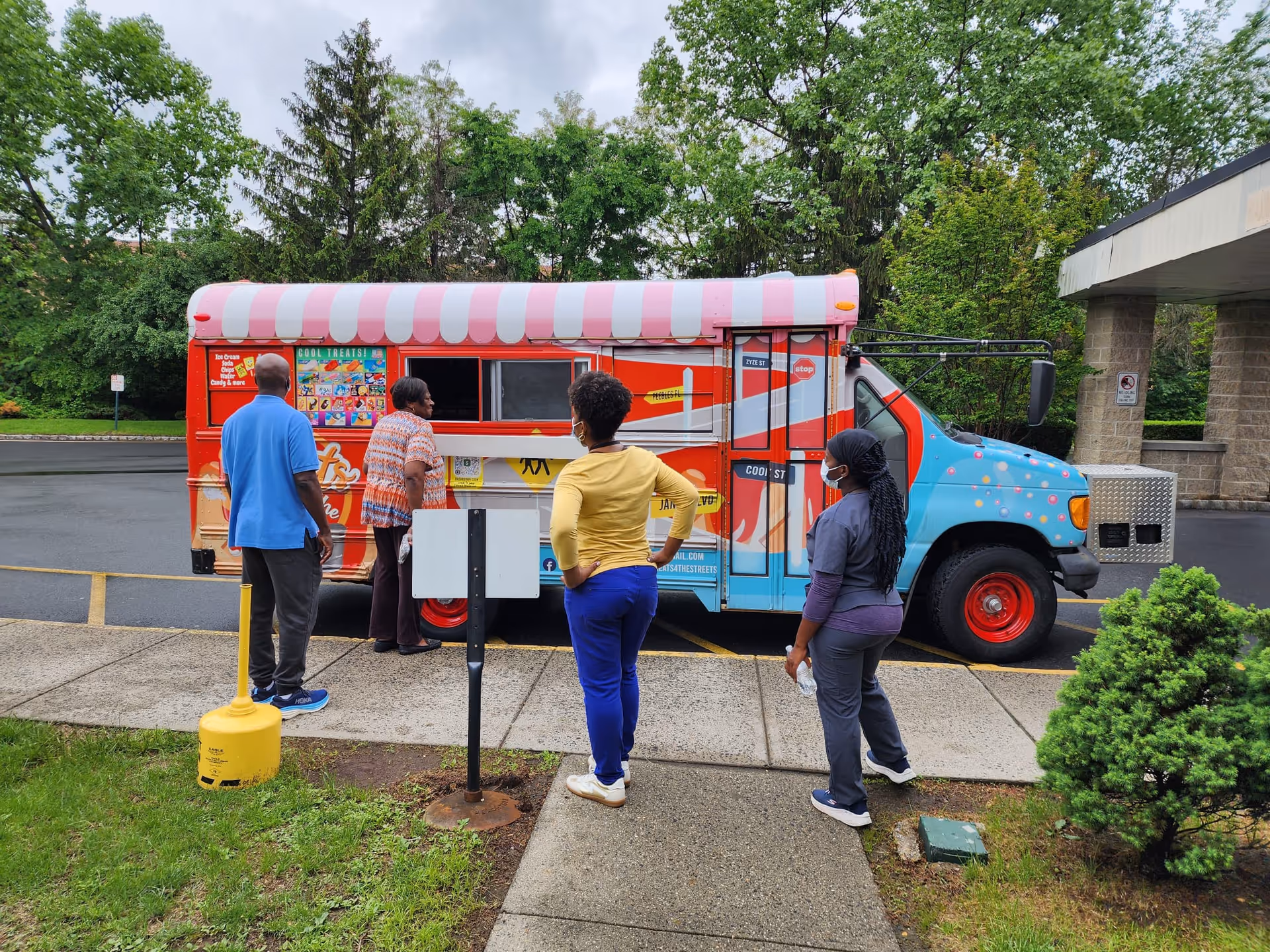Four people standing in line outside a colorful ice cream truck with a pink and white striped awning, parked on a driveway near a building entrance surrounded by greenery.