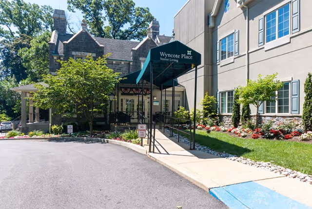Entrance to Wyncote Place Senior Living facility featuring a covered walkway with a green canopy, surrounded by landscaped greenery and flowers, with a paved driveway and a 'No Parking' sign.