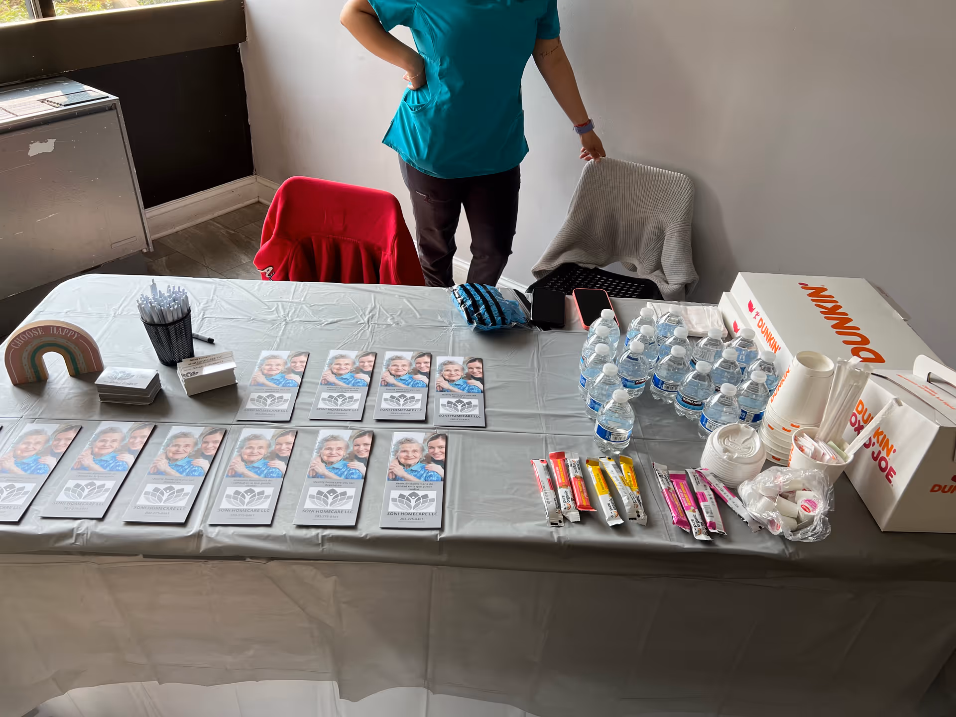 A staff member in teal scrubs stands behind a table of flyers and drinks indoors.