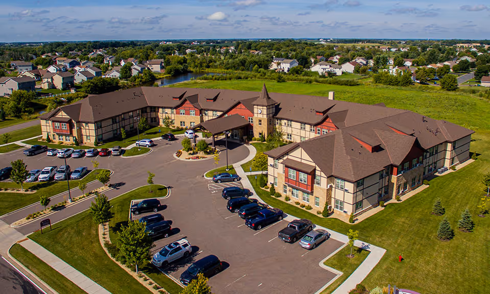 Aerial view of Guardian Angels Engel Haus Senior Living facility in Albertville, showing a large two-story building with a brown roof, multiple parking lots with cars, surrounding green lawns, and a clear blue sky with scattered clouds.