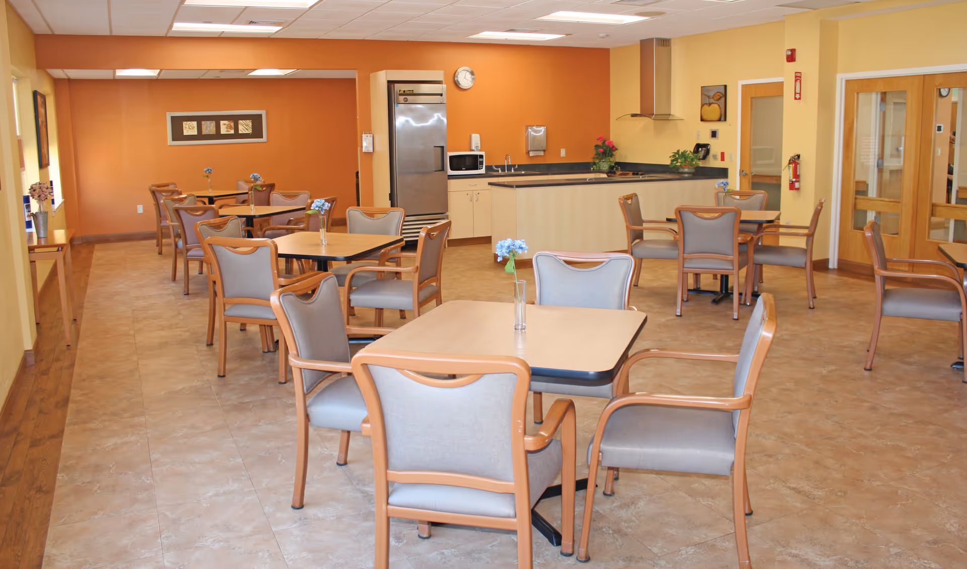 A dining area with several square tables and cushioned chairs arranged neatly. Each table has a small vase with blue flowers. The room has warm yellow and orange walls, a tiled floor, and a kitchen area with a refrigerator, microwave, and sink in the background. There are framed pictures on the walls and a clock above the kitchen area.