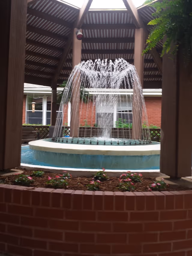 A circular water fountain with water jets creating an arching spray, located under a wooden pavilion roof. The fountain is surrounded by a brick planter with small flowering plants. In the background, there are windows and a brick building wall.