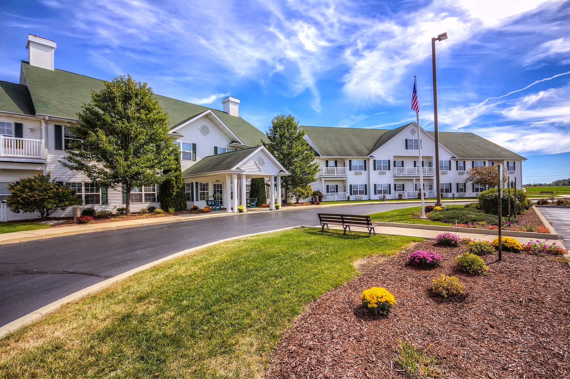 Front exterior of Jefferson Manor senior living facility with a covered entrance, flagpole, bench, driveway and landscaped lawns under a bright sky.