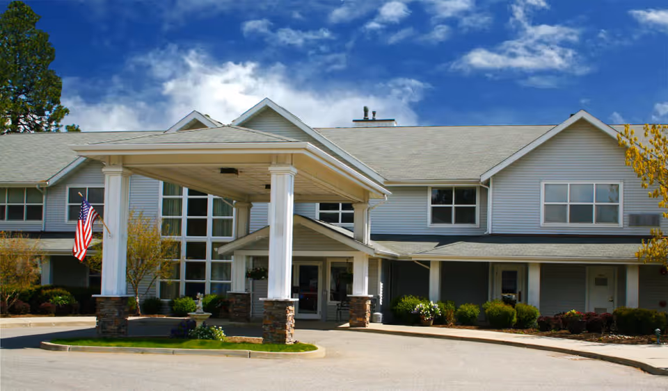 Front exterior of a two-story assisted living building with a covered porte-cochère, columns, American flag, and landscaped entrance under a blue sky.