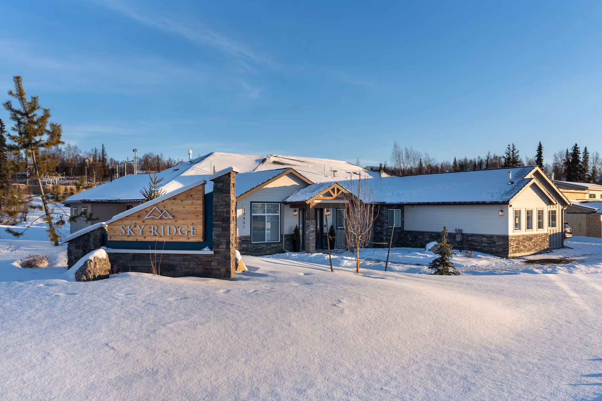 Snow-covered single-story building with a prominent 'SKY RIDGE' entrance sign and a snowy front yard under a clear blue sky.