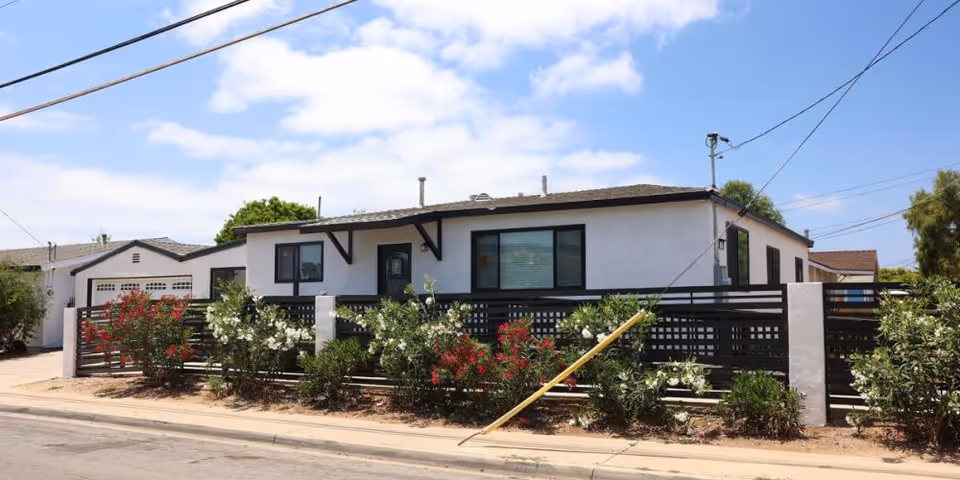 Exterior view of a single-story white building with a black fence and flowering bushes in front, under a partly cloudy blue sky.