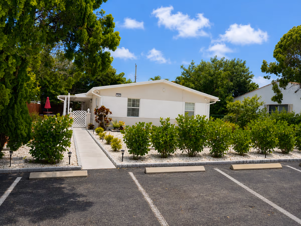 Single-story white building with a paved walkway, shrubs, and a parking lot in foreground.