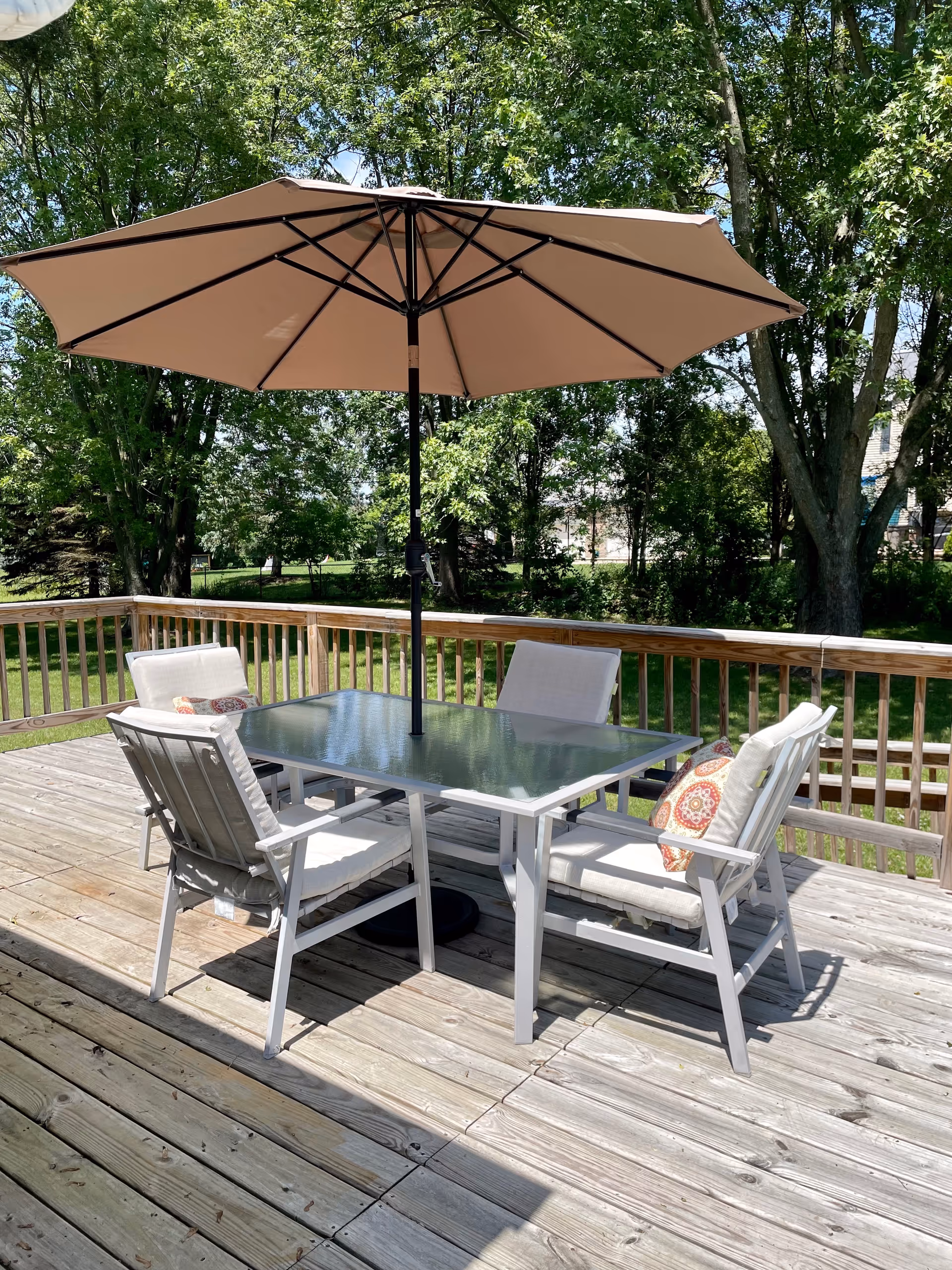 Outdoor wooden deck with glass table, umbrella, and cushioned chairs in sunlight.