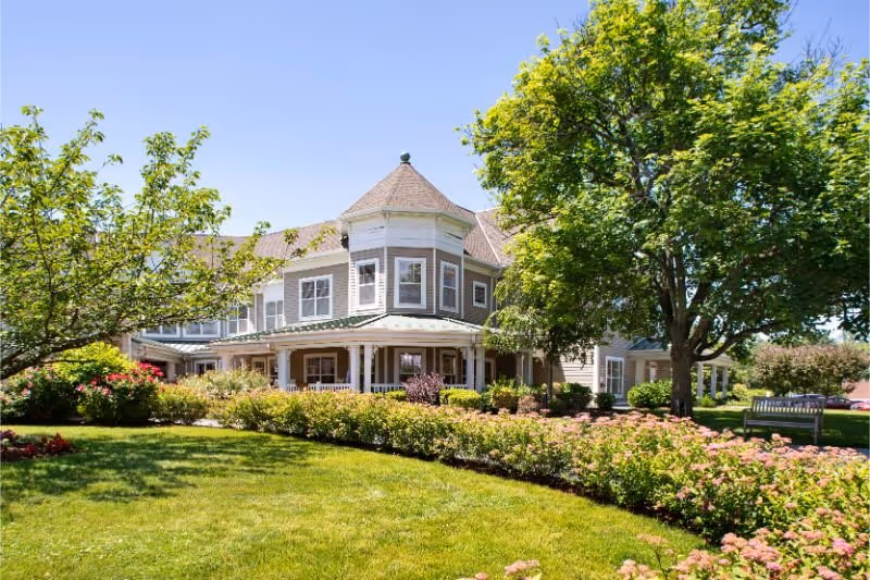 Victorian-style senior living building with a wraparound porch surrounded by a manicured lawn, trees, and flowering shrubs under a clear blue sky.