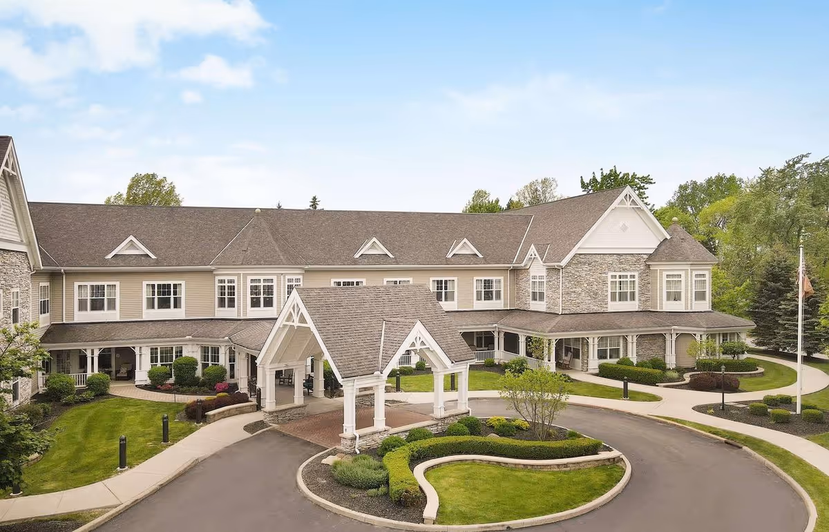 Front exterior of a two-story senior living building with a covered porte-cochere, circular driveway, and landscaped grounds.