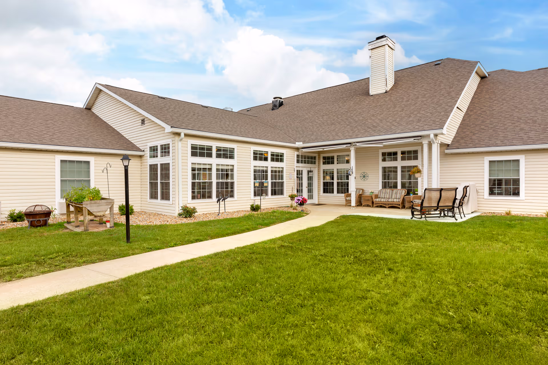 Exterior view of a single-story beige senior living building with large windows, a patio seating area, and a grassy lawn.