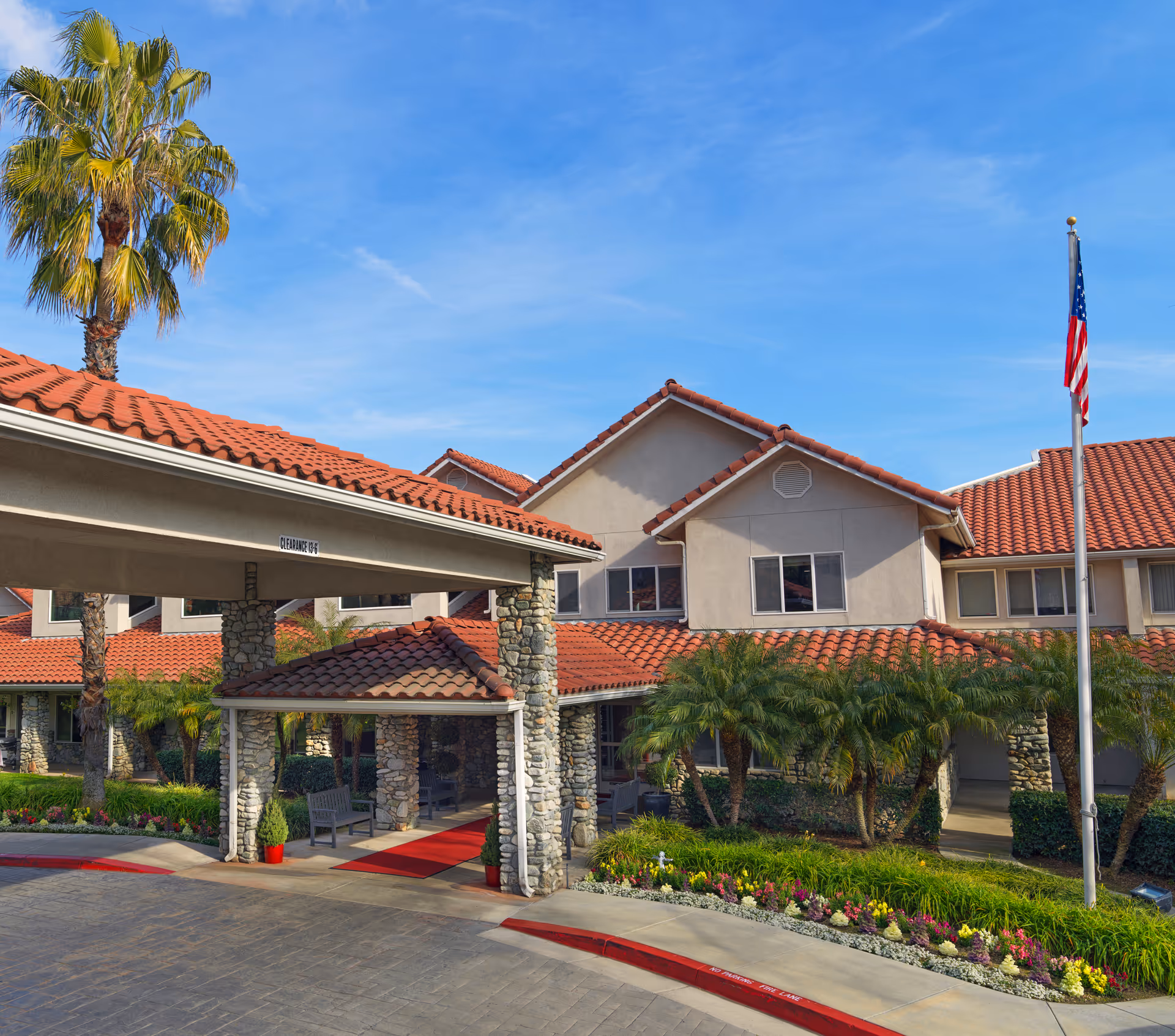 Front entrance of a stucco building with red tile roofs, stone columns, palm trees, and an American flag.