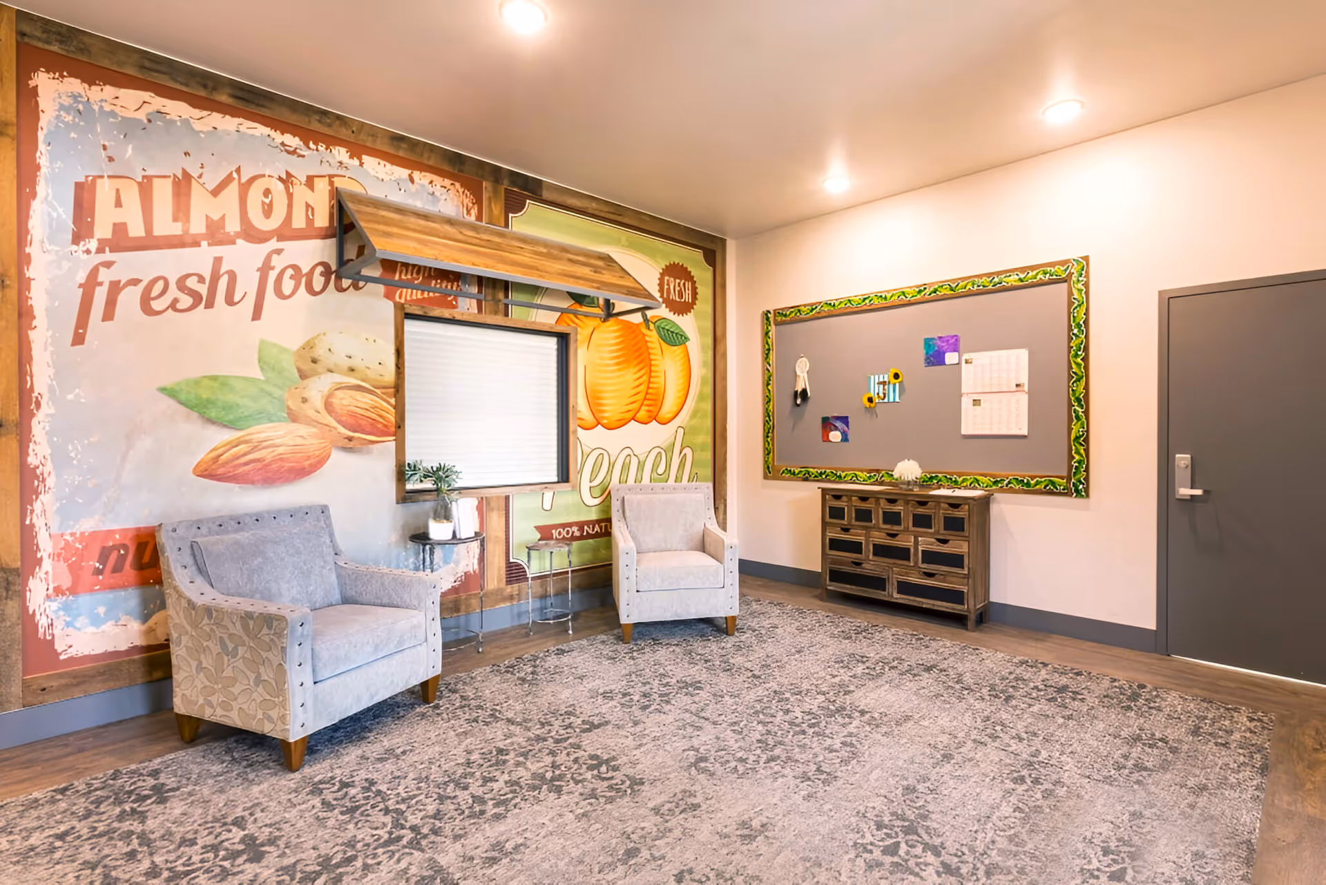 Two light gray upholstered chairs sit beside a fruit-themed mural in a cozy indoor lobby.