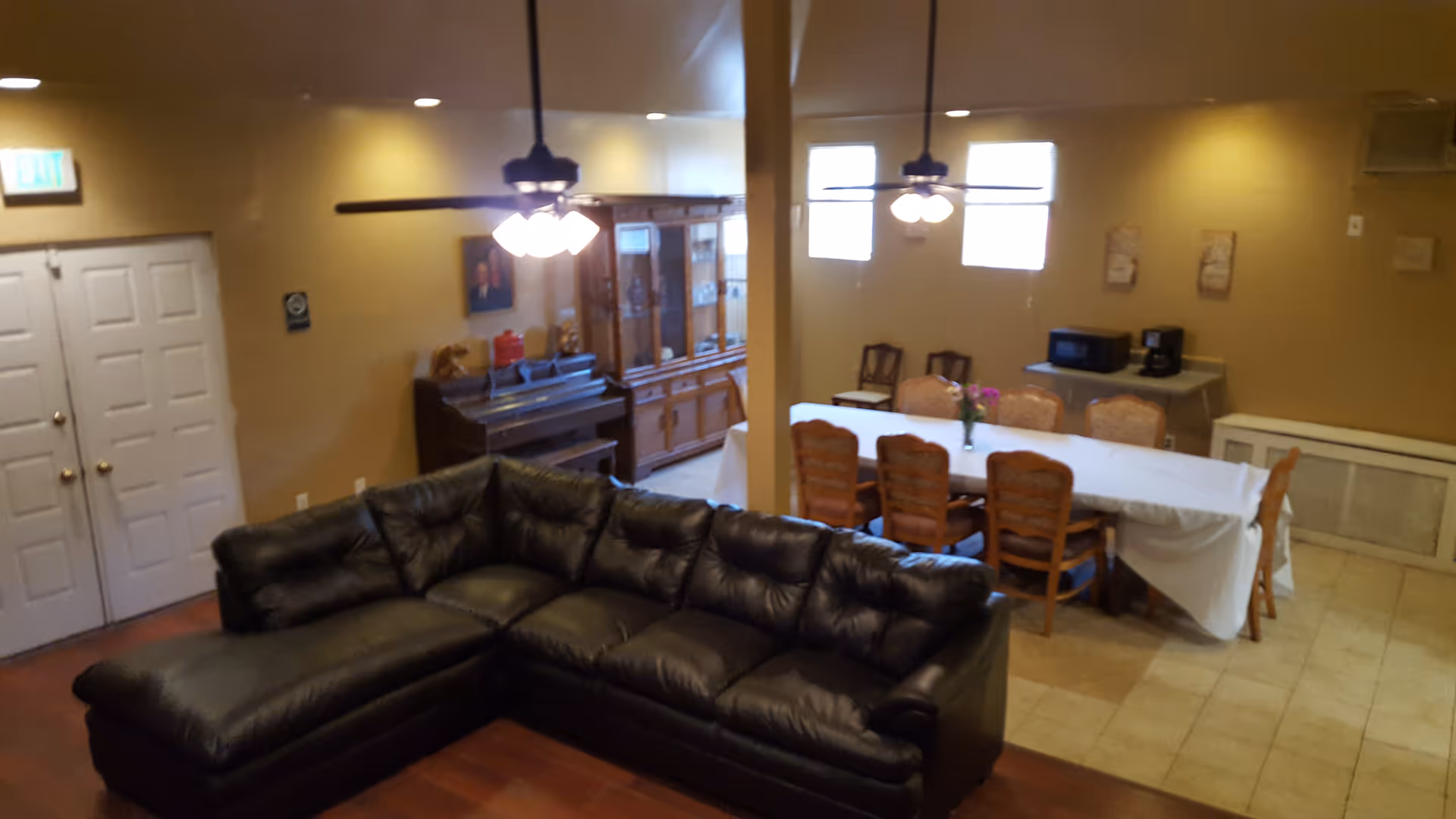 Interior view of a senior living facility room with a black leather sectional sofa in the foreground, a dining table covered with a white tablecloth and surrounded by wooden chairs in the background, ceiling fans with lights, a wooden cabinet, a piano, and a small table with a microwave and coffee maker against the wall.