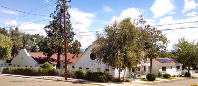 Front exterior view of a single-story white building with a red roof and trees.