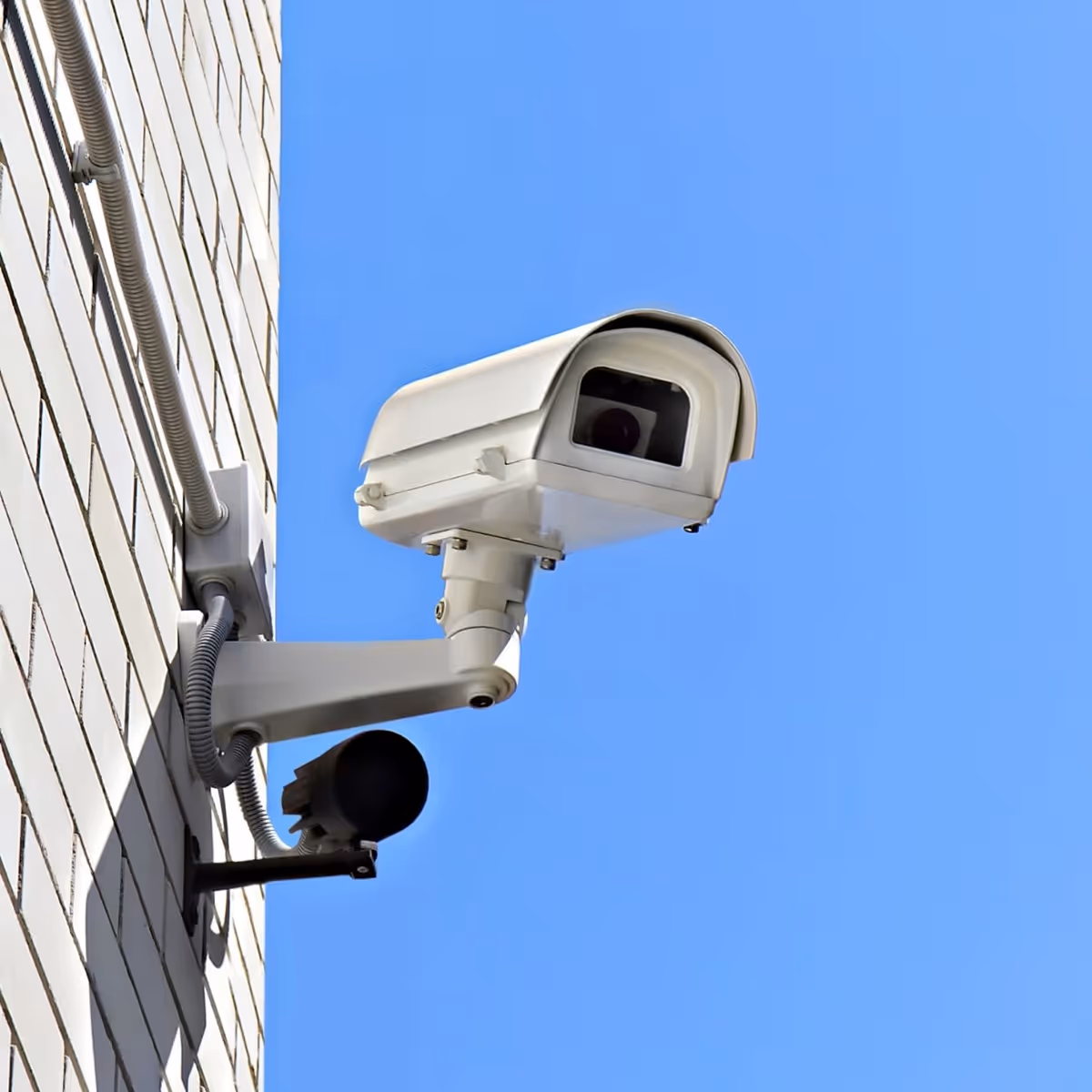 White security camera mounted on building exterior with blue sky background.