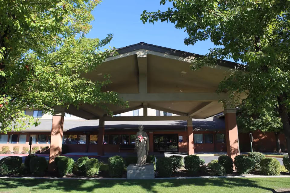 Covered entrance of a building with a porte-cochère, a statue, and landscaped shrubs under trees.