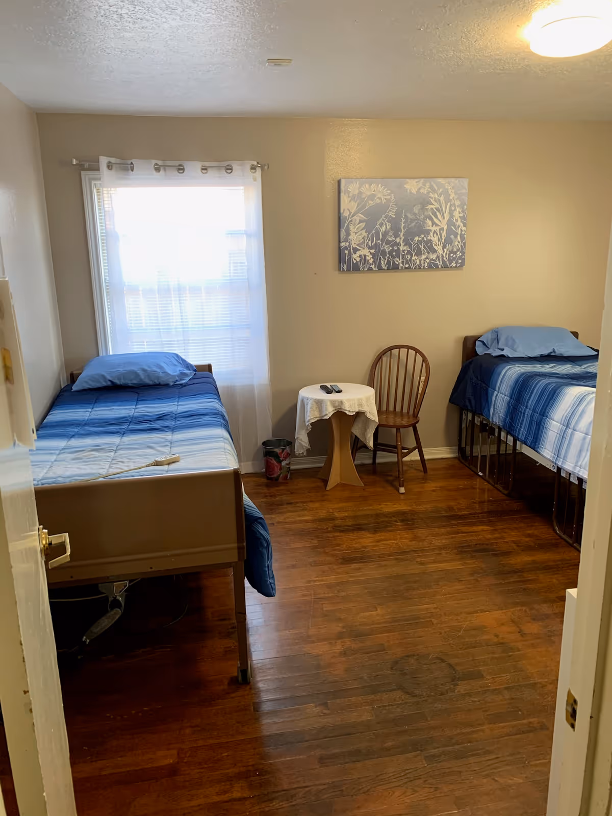 A small shared bedroom with two blue beds, a window, table, and chair.