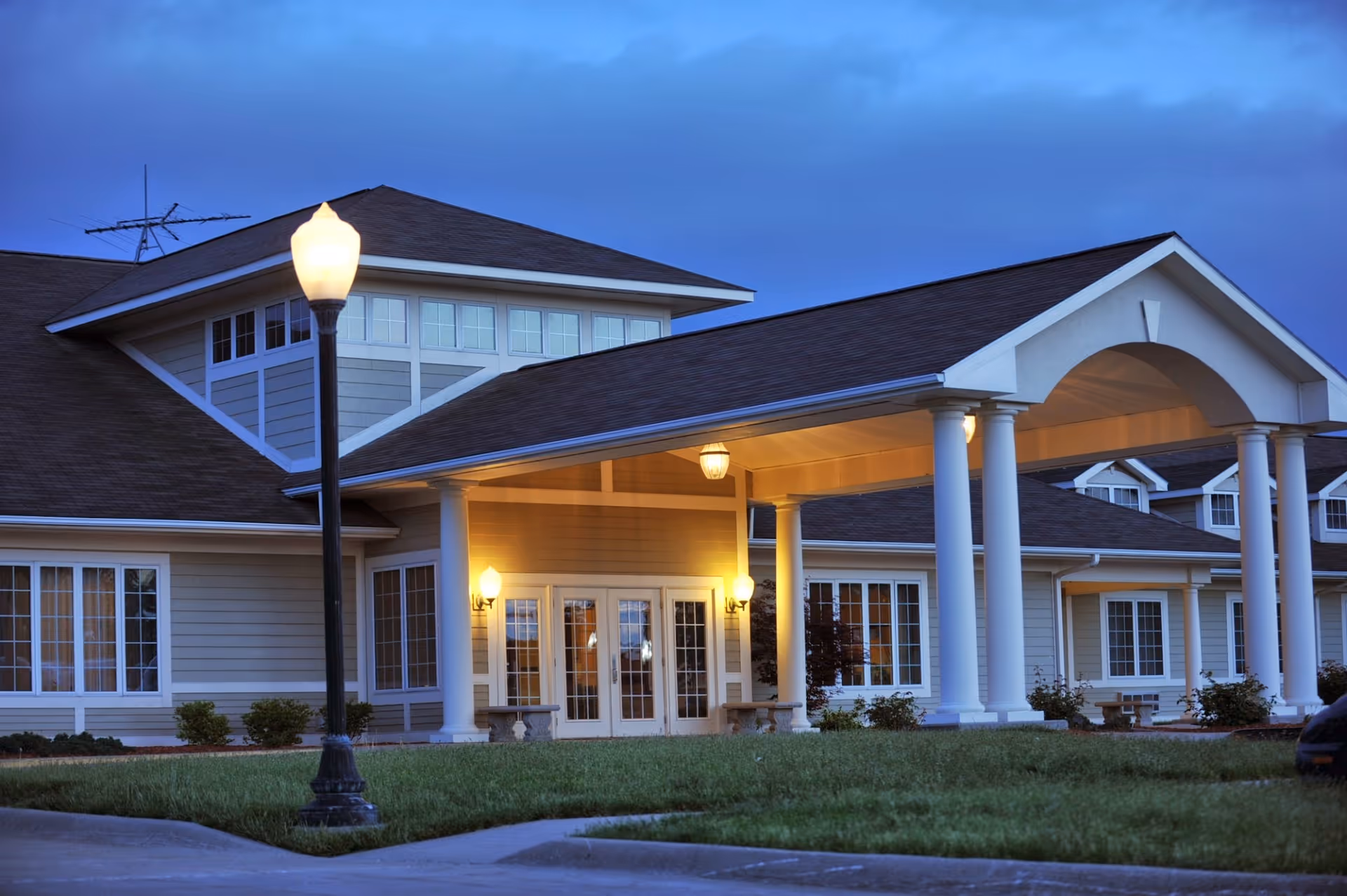 Senior living facility front entrance with columns, lights, and a covered drop-off area at dusk.