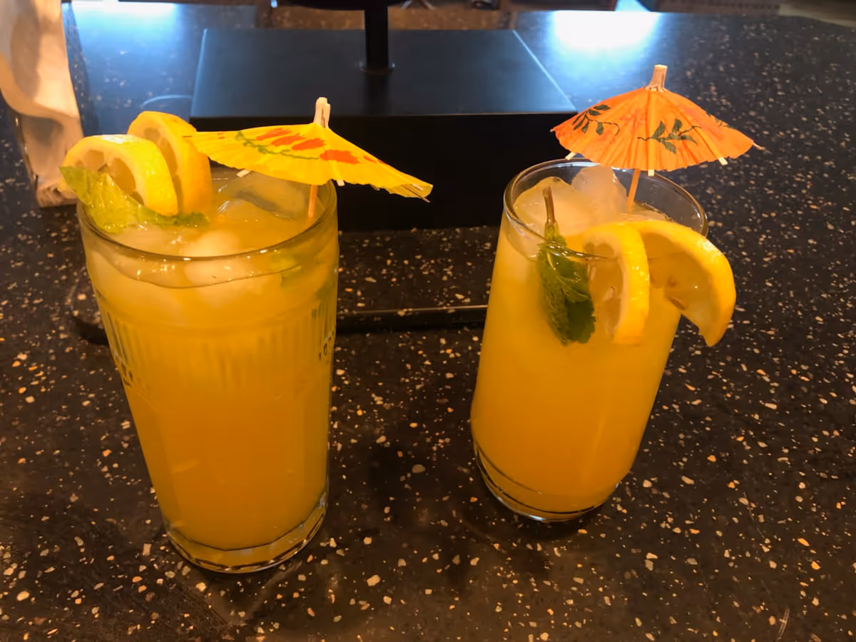 Two tall yellow drinks with lemon slices and paper umbrellas on a speckled bar counter.