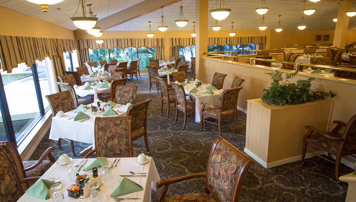 Sunlit senior dining room with white tablecloths, green napkins, and floral upholstered chairs.