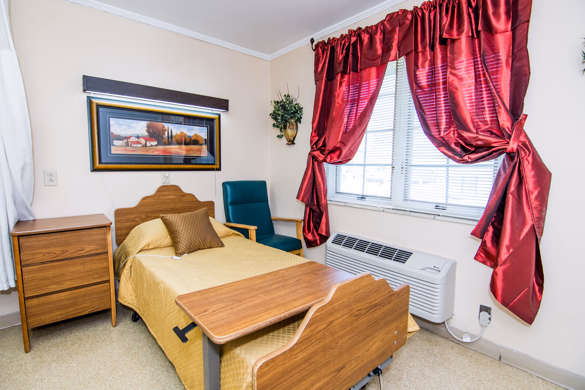 A small, neatly arranged bedroom in a nursing facility featuring a single bed with a wooden headboard and a yellow bedspread, a brown pillow, a wooden bedside table with three drawers, a green armchair, a wooden overbed table, a window with red curtains tied back, and a wall-mounted air conditioning unit below the window. A framed landscape painting hangs above the bed, and a small plant is mounted on the wall.