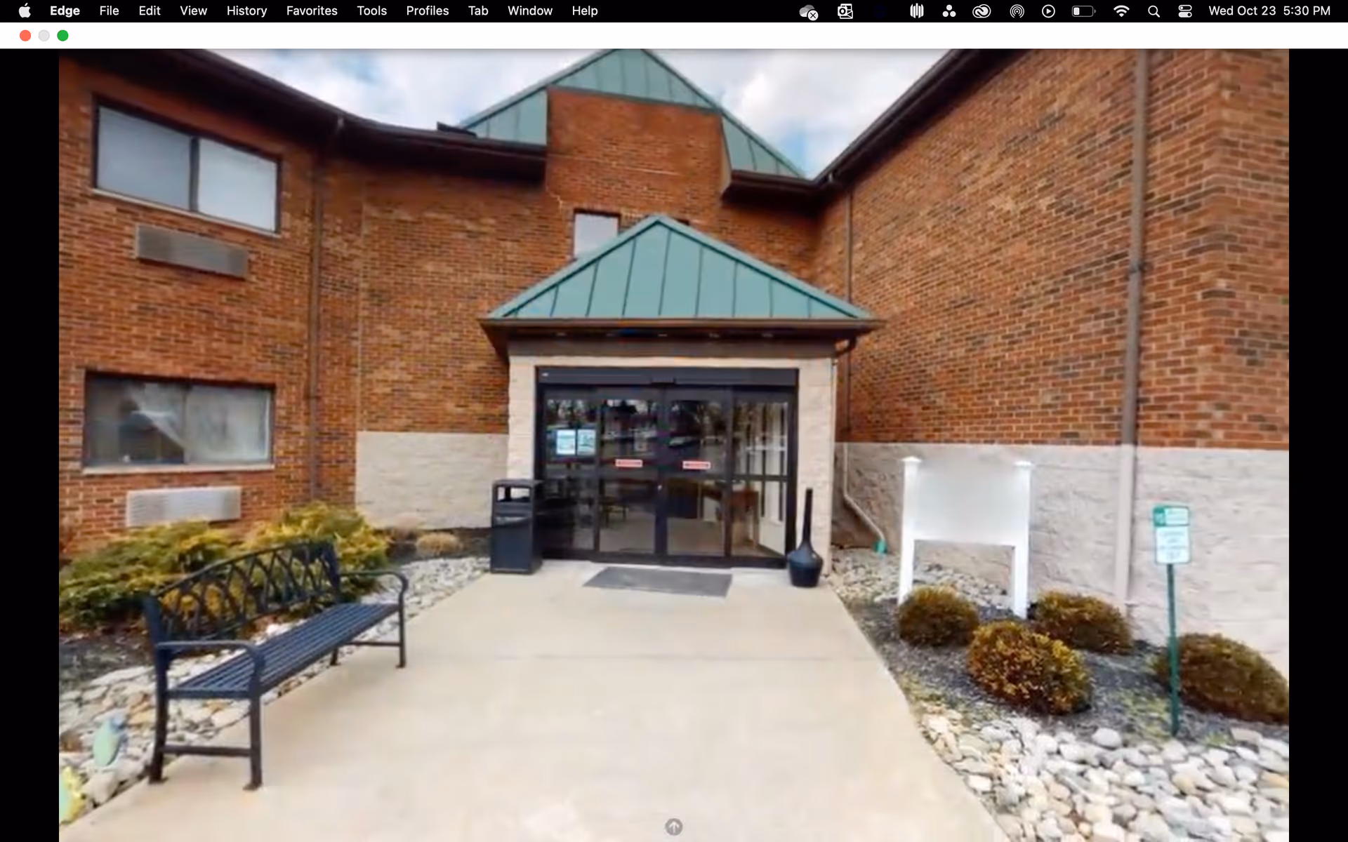 Front entrance of a brick building with a green metal awning over glass double doors, a bench and landscaped walkway.