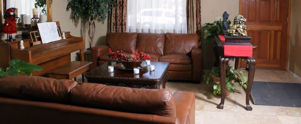 A cozy living room with two brown leather sofas facing a dark wooden coffee table decorated with candles and a centerpiece. There is a wooden piano with sheet music on it to the left, a tall green plant behind the sofa, and a small table with decorative items near the wooden front door. Light filters in through a window with sheer curtains and patterned drapes.